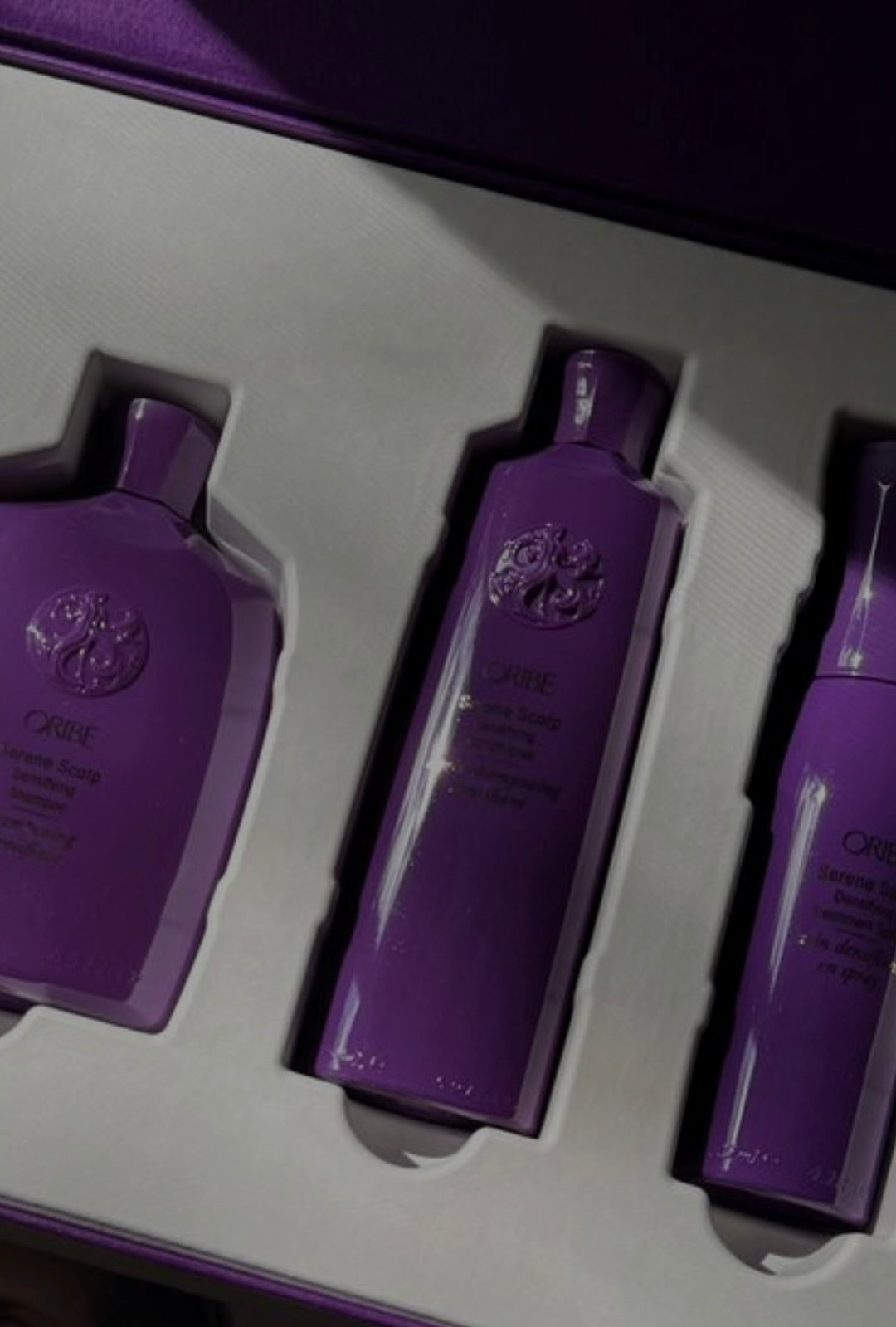 A luxurious set of three Oribe Serene Scalp products in sleek purple bottles displayed in an elegant white and purple gift box, showcasing the brand’s signature embossed design and premium packaging.