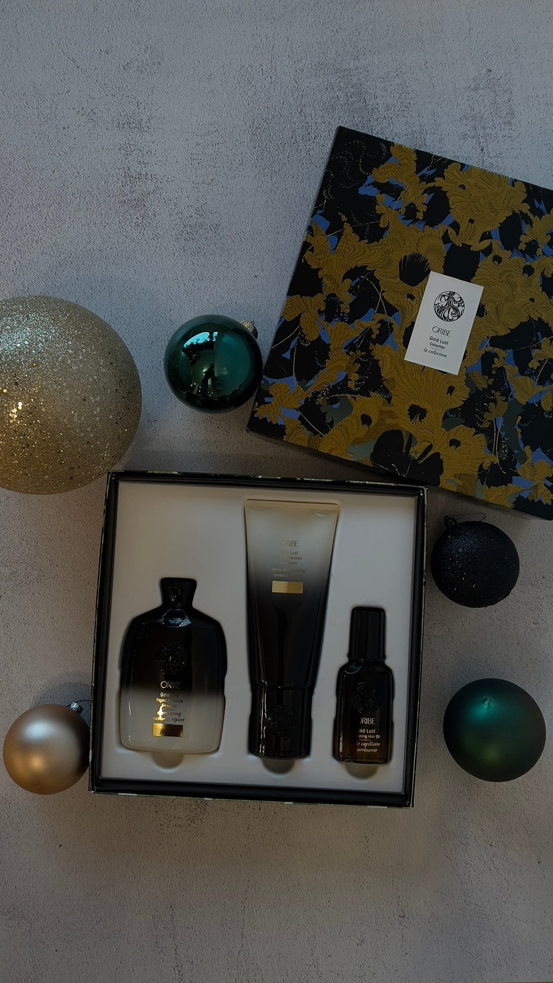 Oribe Gold Lust holiday gift set displayed in an open box surrounded by Christmas ornaments.