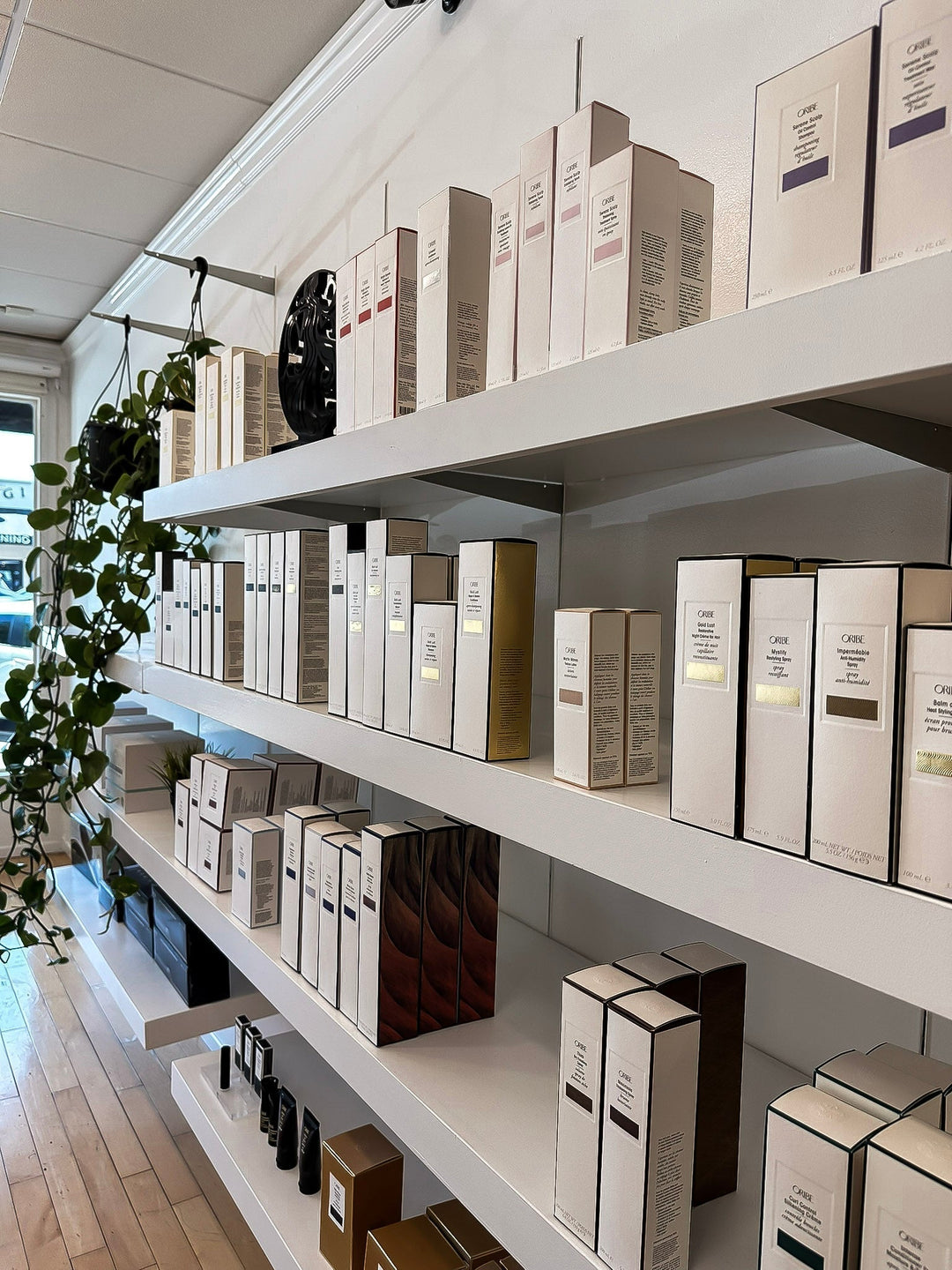 Shelves filled with neatly arranged Oribe haircare products in elegant cream, gold, and bronze packaging, displayed in a modern salon retail area with natural light and hanging greenery.