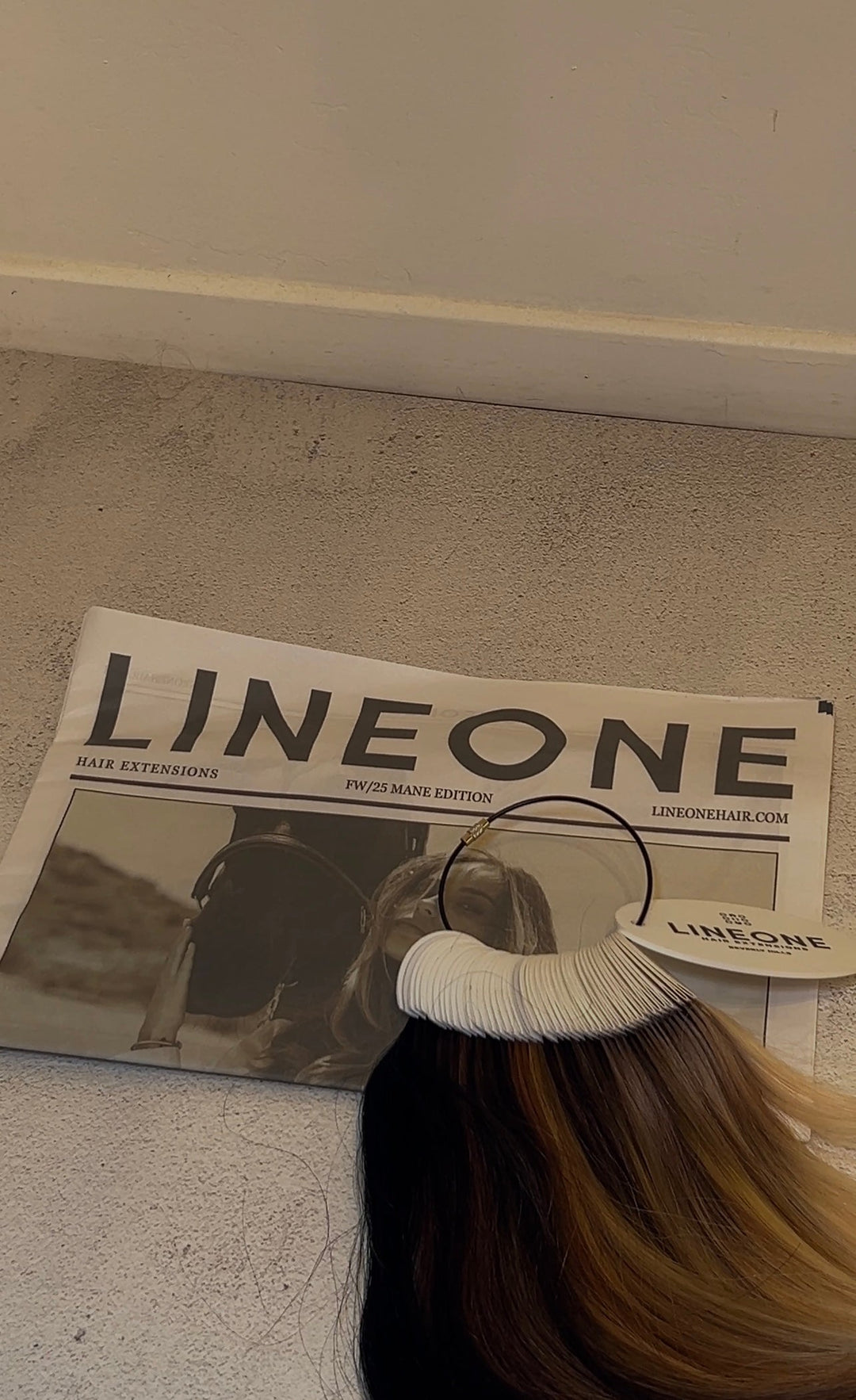 Line One Hair Extensions display with multi-tonal hair weft on hanger against brand magazine backdrop