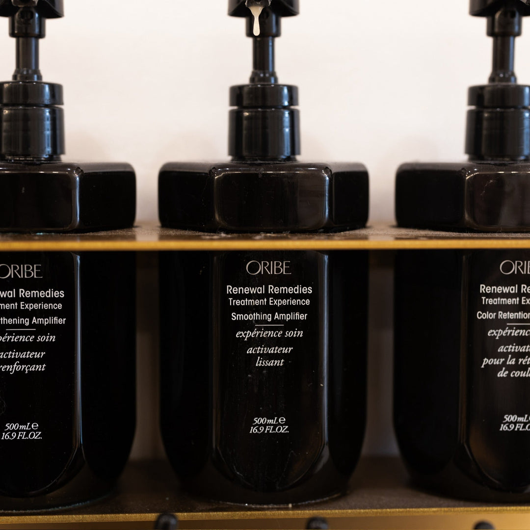 Close-up of black Oribe Renewal Remedies Treatment Experience bottles displayed on a salon shelf, showcasing the Smoothing Amplifier formula in sleek, minimalist packaging with gold accents.