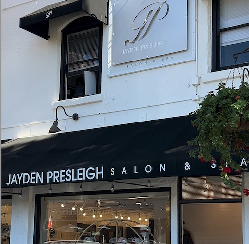 Exterior view of Jayden Presleigh Salon & Spa featuring a white stucco building with black awnings, a hanging metal sign with the salon’s logo, and large windows revealing the elegant interior and salon chairs inside.