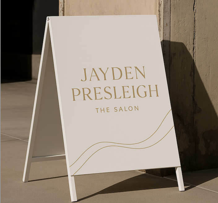 Elegant cream-colored sandwich board sign with gold lettering that reads “Jayden Presleigh The Salon,” featuring a minimal wavy line design at the bottom, positioned outdoors against a textured wall in natural light.