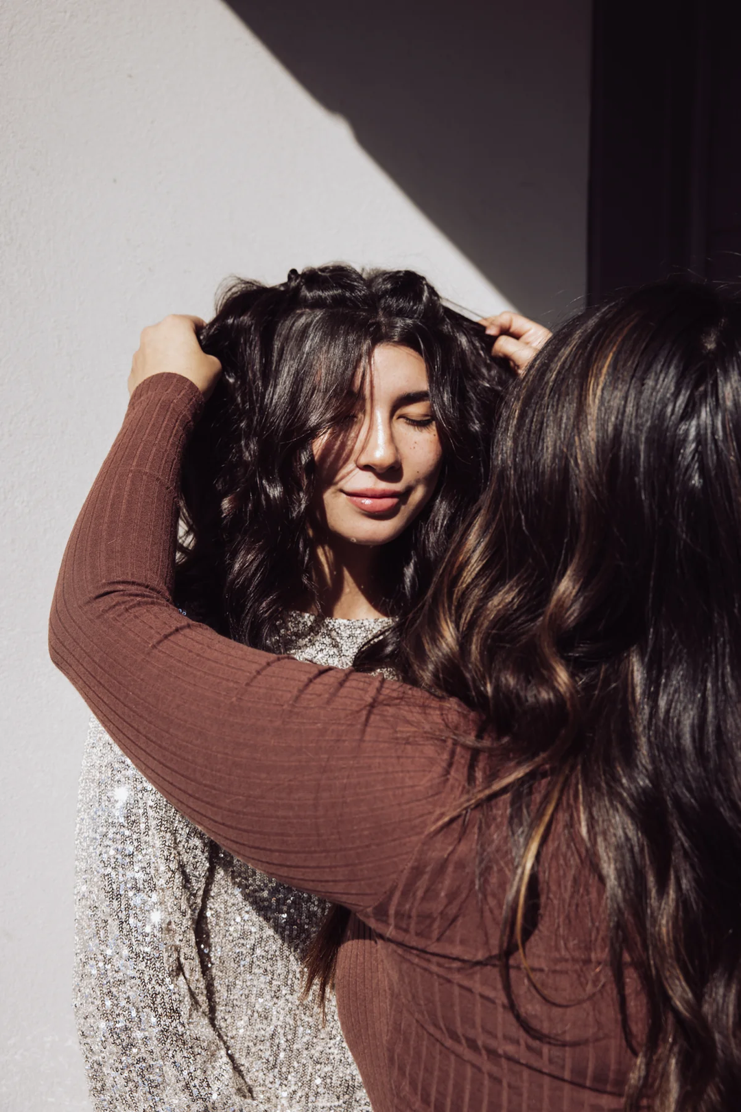 Hairstylist adjusting a client’s loose, wavy curls in natural sunlight. The client, wearing a sparkly silver top, smiles softly with her eyes closed against a neutral wall.